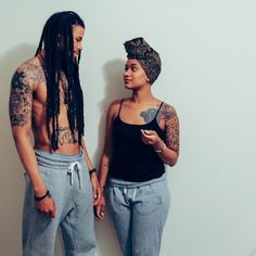 two people standing next to each other in front of a wall with tattoos on their arms