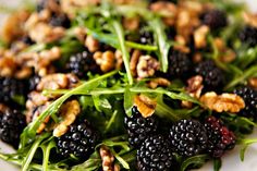 a white plate topped with blackberries and green leafy salad next to walnuts