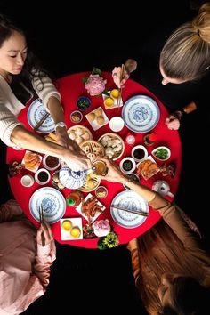 several people sitting around a red table with food on it and plates in the middle