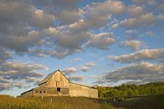 an old barn sits in the middle of a grassy field with trees and clouds above it