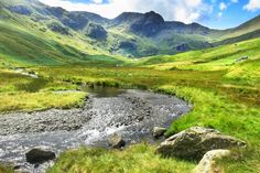 a stream running through a lush green valley filled with mountains in the distance, surrounded by grass and rocks