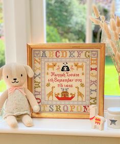 a teddy bear sitting next to a framed cross - stitch sample on a window sill