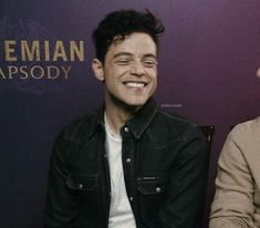 a smiling young man sitting in front of a purple wall with the words bohemian apsody on it