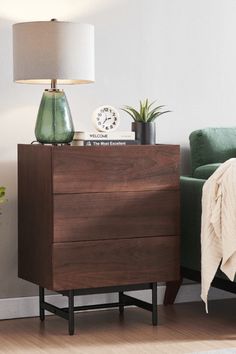 a night stand with a lamp on it next to a green chair and a plant