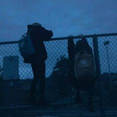 two people standing next to each other with backpacks on their backs and one person leaning against a fence