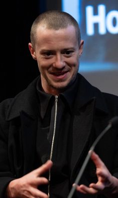 a man with a shaved head and black shirt is talking into a microphone while holding his hands out in front of him