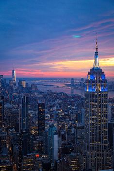 the empire state building lit up in blue and white at dusk, as seen from top of the rock