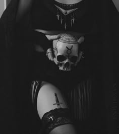 a woman in black clothing holding a skull and cross tattoo on her arm while sitting down