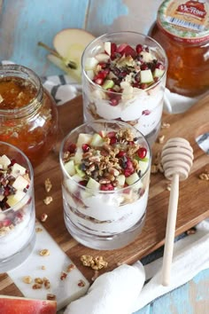 three glasses filled with dessert sitting on top of a cutting board next to apples and honey