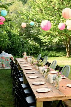 a long wooden table topped with lots of plates and cups filled with flowers next to paper lanterns