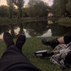 two people sitting next to each other on the grass near a lake with trees and mountains in the background