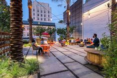 people are sitting at tables and chairs in an outdoor patio area with palm trees, potted plants, and hanging lights