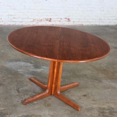 a round wooden table with four legs on concrete flooring in front of a brick wall