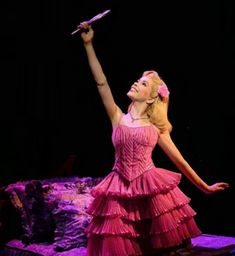 a woman in a pink dress is performing on stage with her arms up and hands out