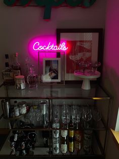 a bar cart filled with liquor bottles and wine glasses next to a neon sign that reads cocktails