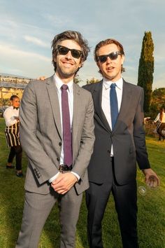 two men standing next to each other in suits and sunglasses on the grass with people behind them