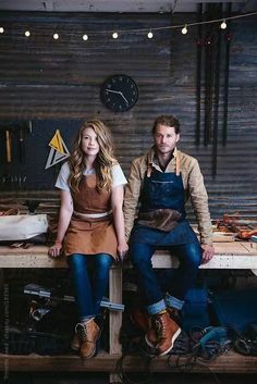 two people are sitting on a bench in a workshop, one is wearing an apron