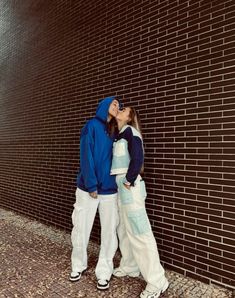 two people standing next to each other in front of a brick wall with one kissing the other