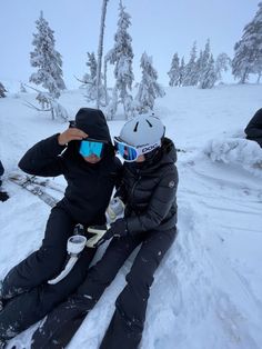 two people sitting in the snow with skis on their feet and one person wearing goggles