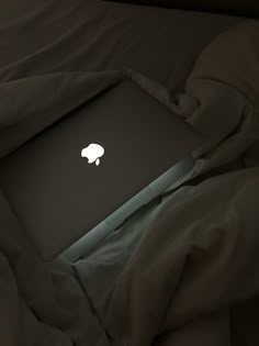 an apple laptop sitting on top of a bed covered in blankets and sheets, with the screen lit up