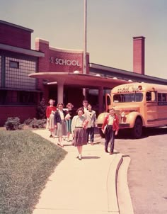 several children are standing in front of a school building with a yellow bus parked next to it