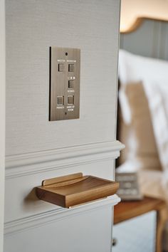 a light switch on the side of a white wall next to a night stand with a bed in the background