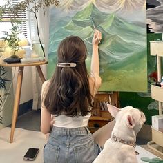 a woman is painting on an easel with a dog sitting next to her in front of the easel