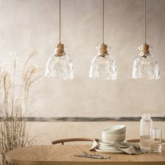 three clear glass pendant lights hanging over a wooden table with plates and utensils