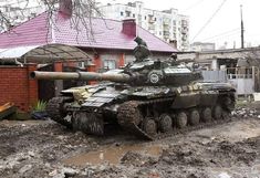 an army tank sitting on top of a muddy road next to a red brick building