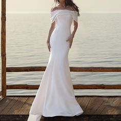 a woman in a white wedding dress standing on a wooden dock near the ocean with her hands on her hips