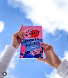 two hands holding up a book against a blue sky with clouds in the back ground