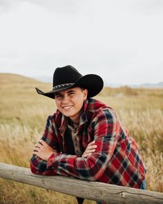 a smiling woman wearing a cowboy hat leaning on a wooden fence in a grassy field