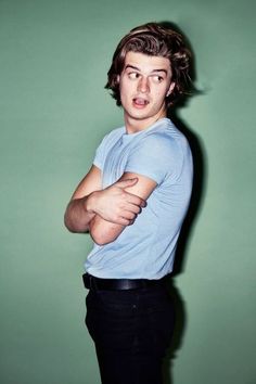 a young man with his arms crossed standing in front of a green wall wearing a blue shirt and black pants