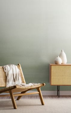 a wooden chair sitting next to a white vase on top of a table in front of a green wall