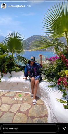 a woman sitting on top of a stone walkway next to flowers and trees with the ocean in the background