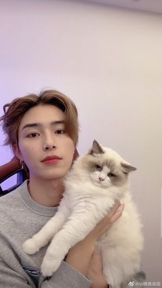 a man holding a white cat in his arms and looking at the camera while wearing a gray shirt