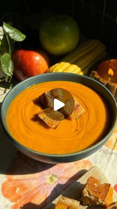 a bowl of carrot soup with crackers and apples in the background