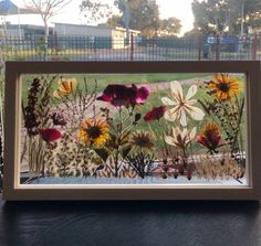 an image of flowers in a frame on a table with grass and fence behind it