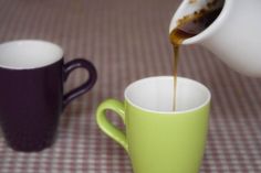 coffee being poured into two mugs sitting on a checkered tablecloth covered table