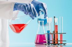 a person in white lab coat and blue gloves pouring red liquid into beakle with test tubes