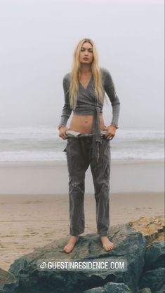 a woman standing on top of a rock next to the ocean with her hands in her pockets