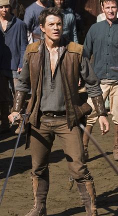 the actor is walking with two swords in his hand and other people are standing behind him