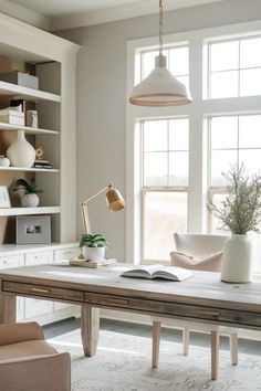 Sunlit home office space with a wooden desk, white chair, pendant light, and book open on the table, suggesting a calm work environment. Transitional Desk Office Spaces, Studio Mcgee Spec Home Office, Feminine Home Office White Desk, Feminine Office Wood Desk, French Partners Desk Home Office, Neutral Home Office, Modern Home Office For Women, Feminine Home Office Classy, Feminine Home Office Ideas