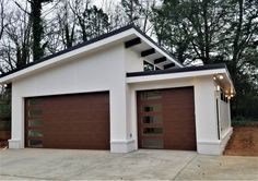 two garages are shown in front of some trees and bushes, one is brown and the other is white