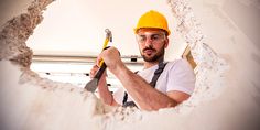 a man in a hardhat holding a hammer and looking through a hole in the wall