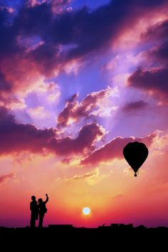 two people standing on top of a hill under a purple sky with a hot air balloon in the distance