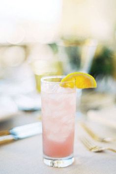 a pink drink with an orange slice on the rim sits on a table next to silverware