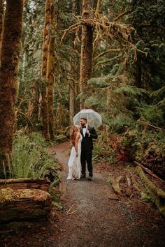 a bride and groom standing under an umbrella in the woods on a path surrounded by trees