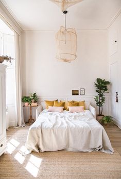 a white bed with yellow pillows in a bedroom next to a window and potted plants