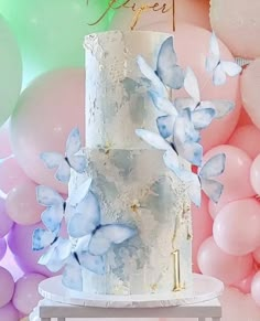 a three tiered cake with blue flowers on it and balloons in the back ground